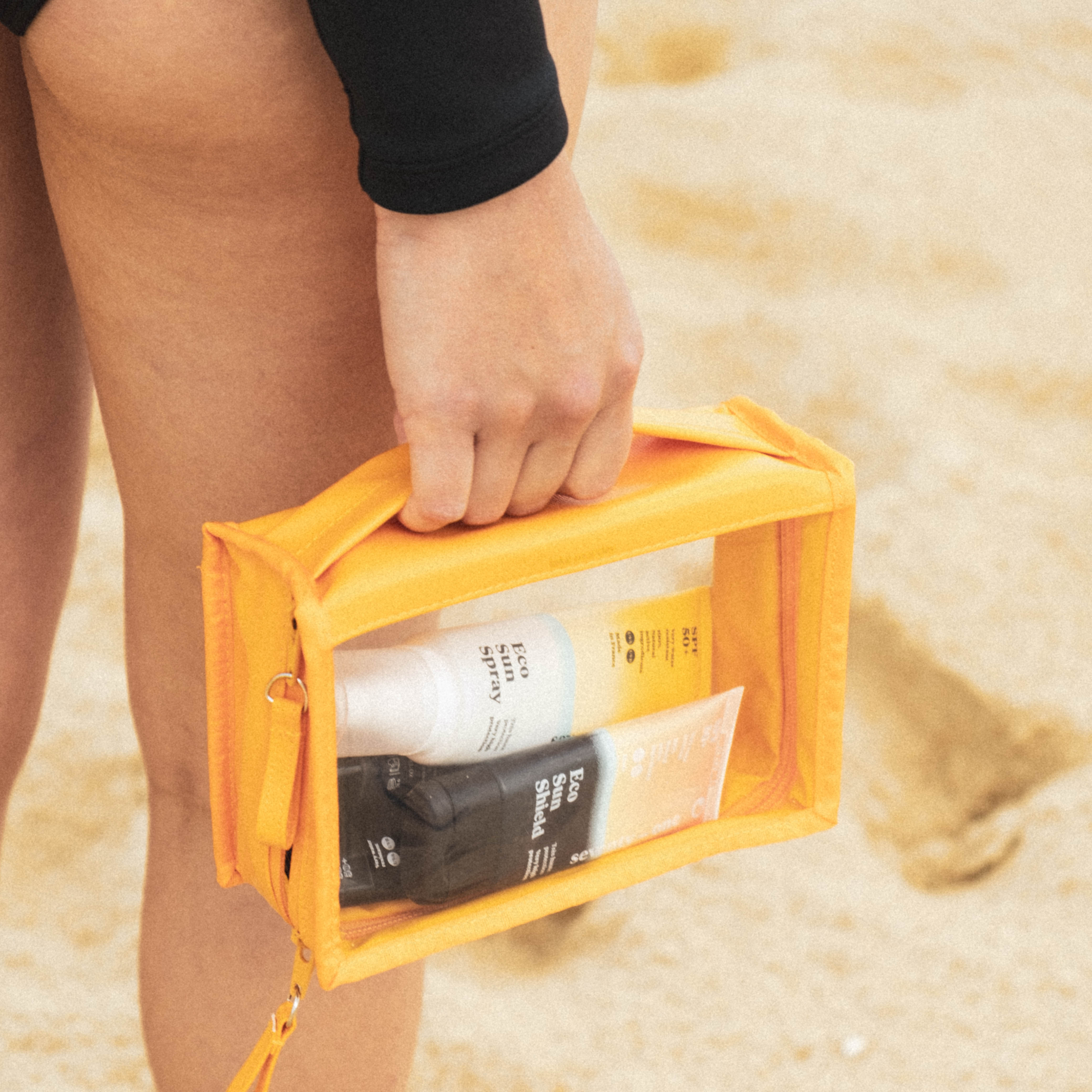 A person's hand holds a bright orange transparent travel bag filled with Eco Sun Spray and Eco Sun Shield bottles on a sandy beach