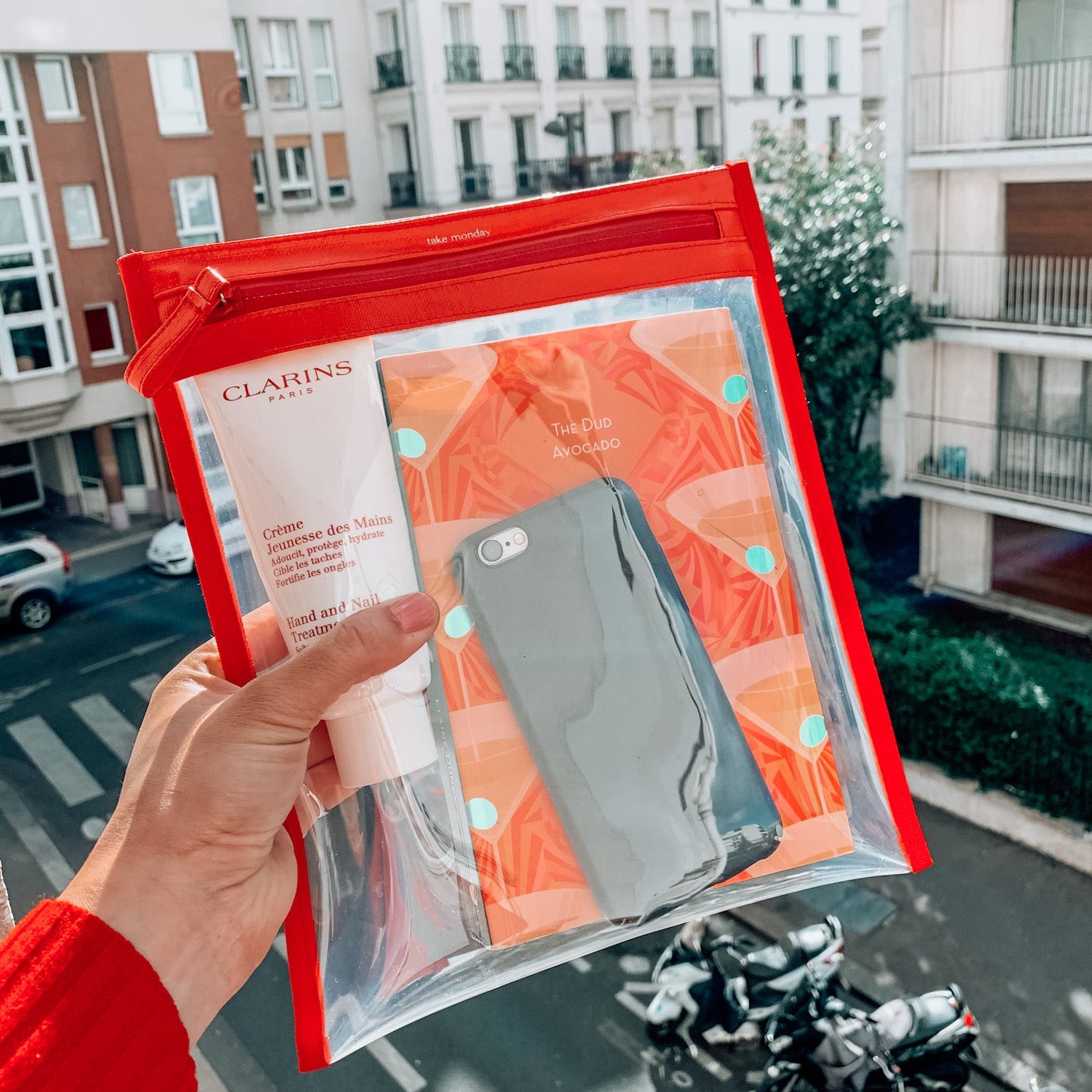 A hand in a red sweater holds a clear red-trimmed "take monday" travel pouch with Clarins hand cream, a grey smartphone, and an orange "The Dud Avocado" book, overlooking a Paris street with Haussmannian buildings and mopeds