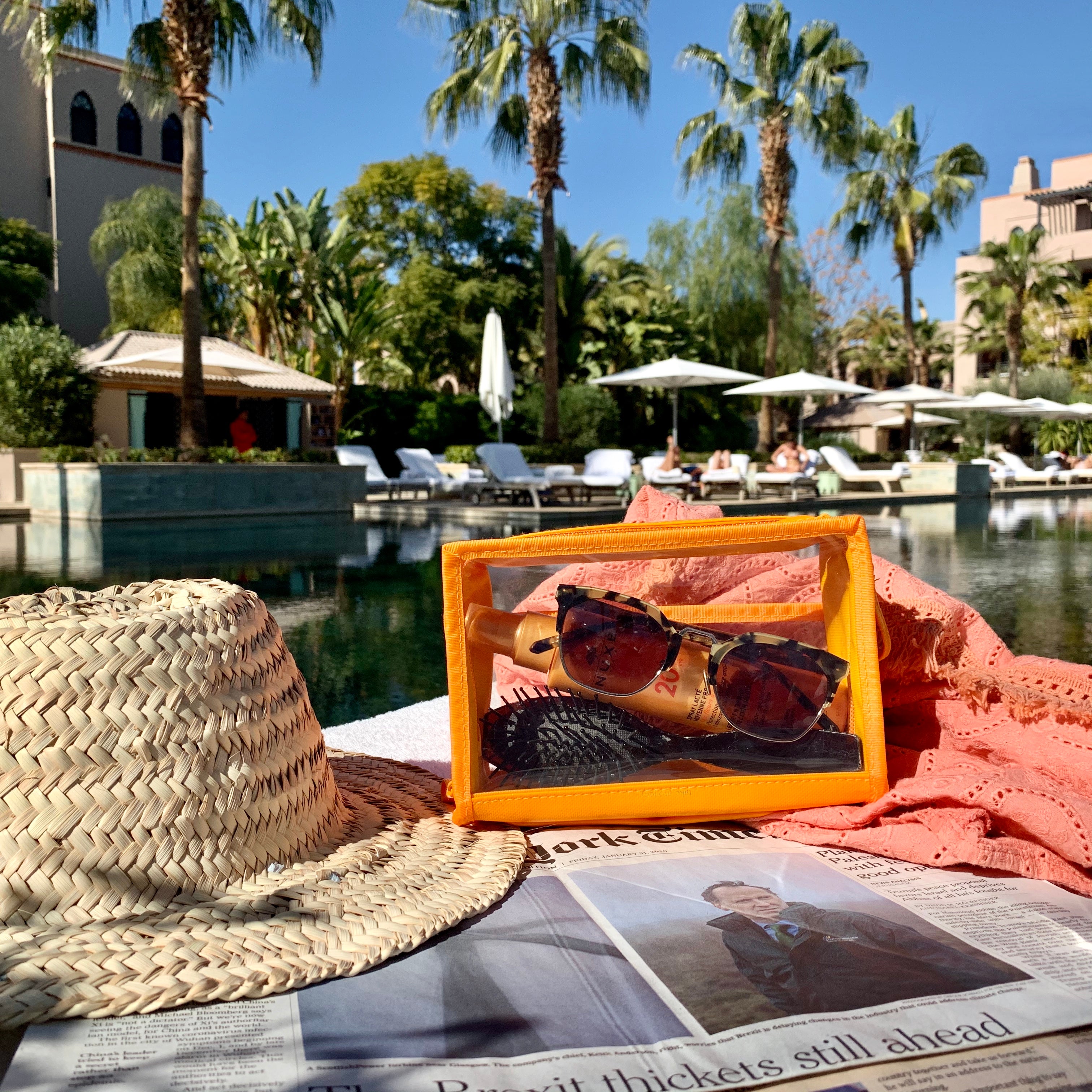 Photograph of a straw hat, a New York Times newspaper, and an orange transparent pouch with tortoiseshell sunglasses and sunscreen resting poolside at a lush resort featuring palm trees and Moorish-style architecture