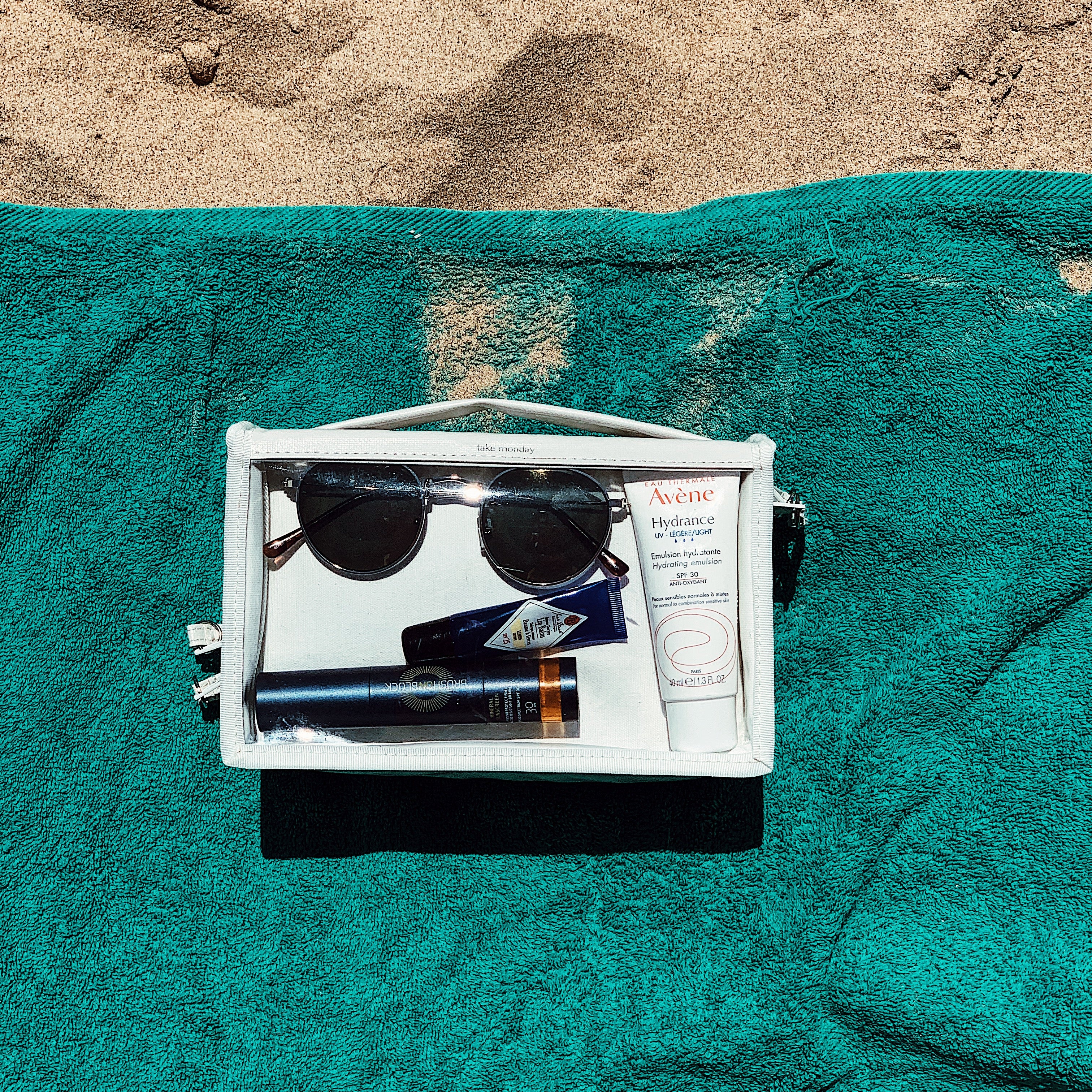 Photography of a white "take monday" transparent cosmetic bag with dark round sunglasses, Avène Hydrance SPF 30, and Brush on Block mineral sunscreen, resting on a vibrant teal beach towel and golden sand