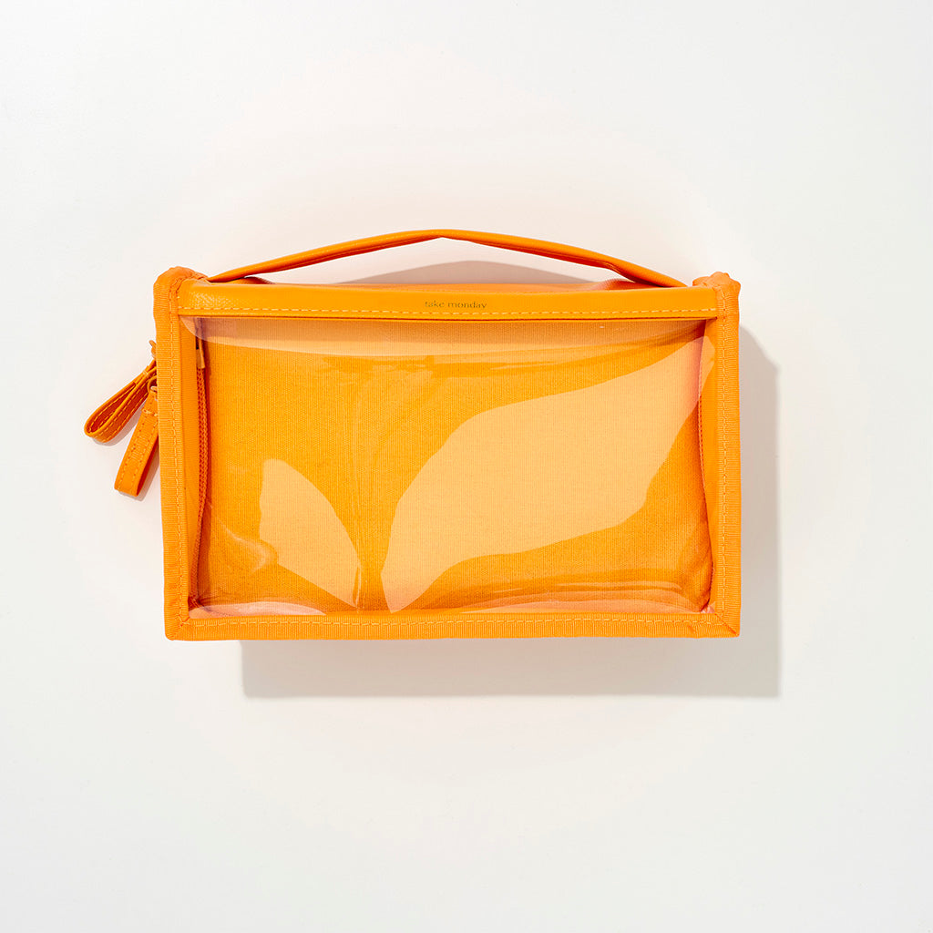 Photograph of a vibrant orange take monday clear-front travel bag featuring a top handle and side zipper on a clean white background