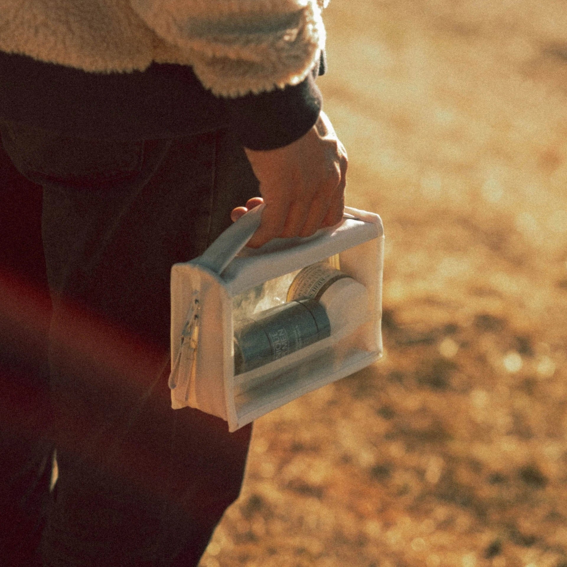 Photography of a person's hand in a fuzzy beige jacket sleeve holding a transparent white toiletry bag filled with travel-sized men's grooming products, bathed in warm golden sunlight outdoors