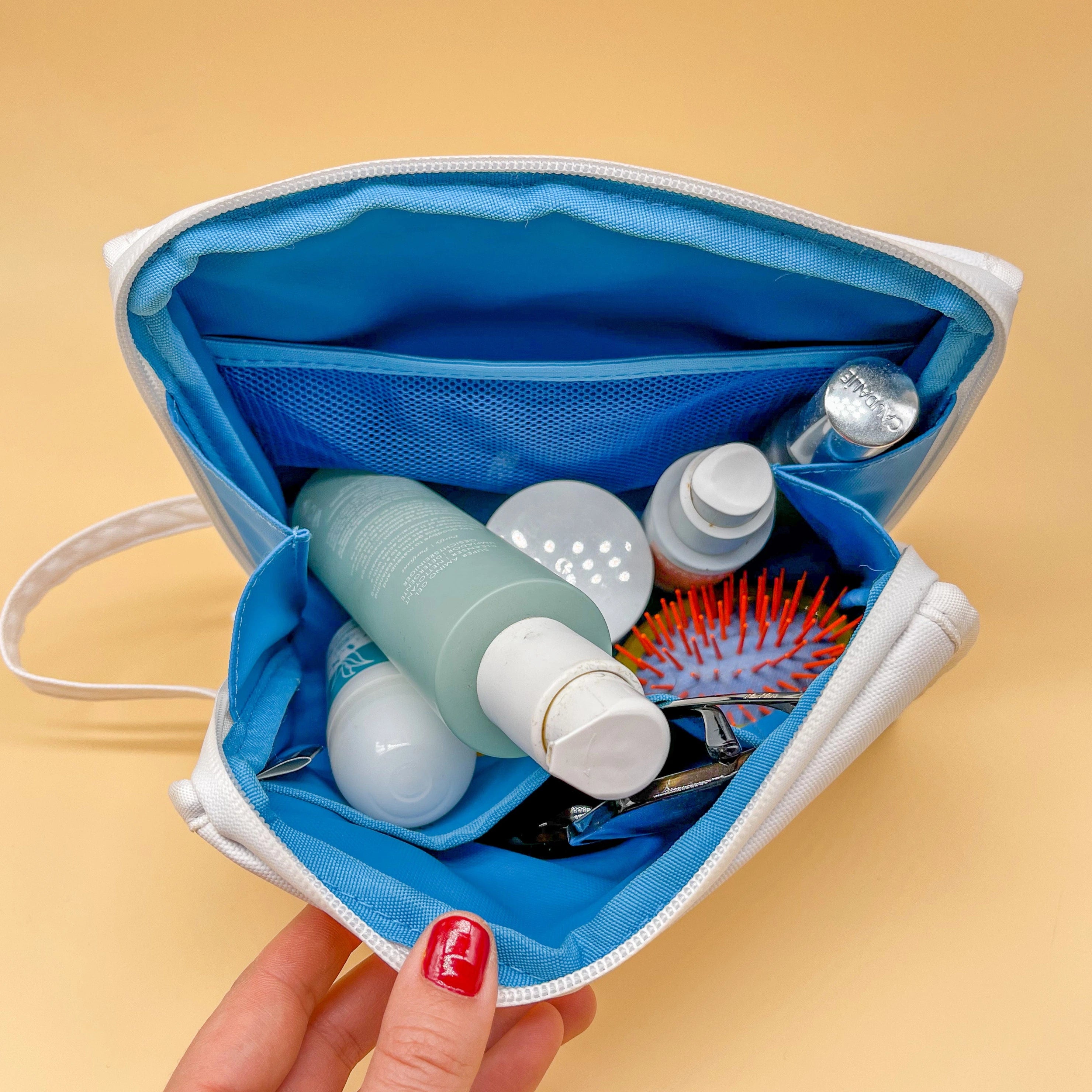 An open white toiletry bag with a bright blue interior held by a hand with red nail polish filled with beauty products including a greenish-blue Youth to the People cleanser bottle a silver Caudalie cap and a vibrant orange hairbrush