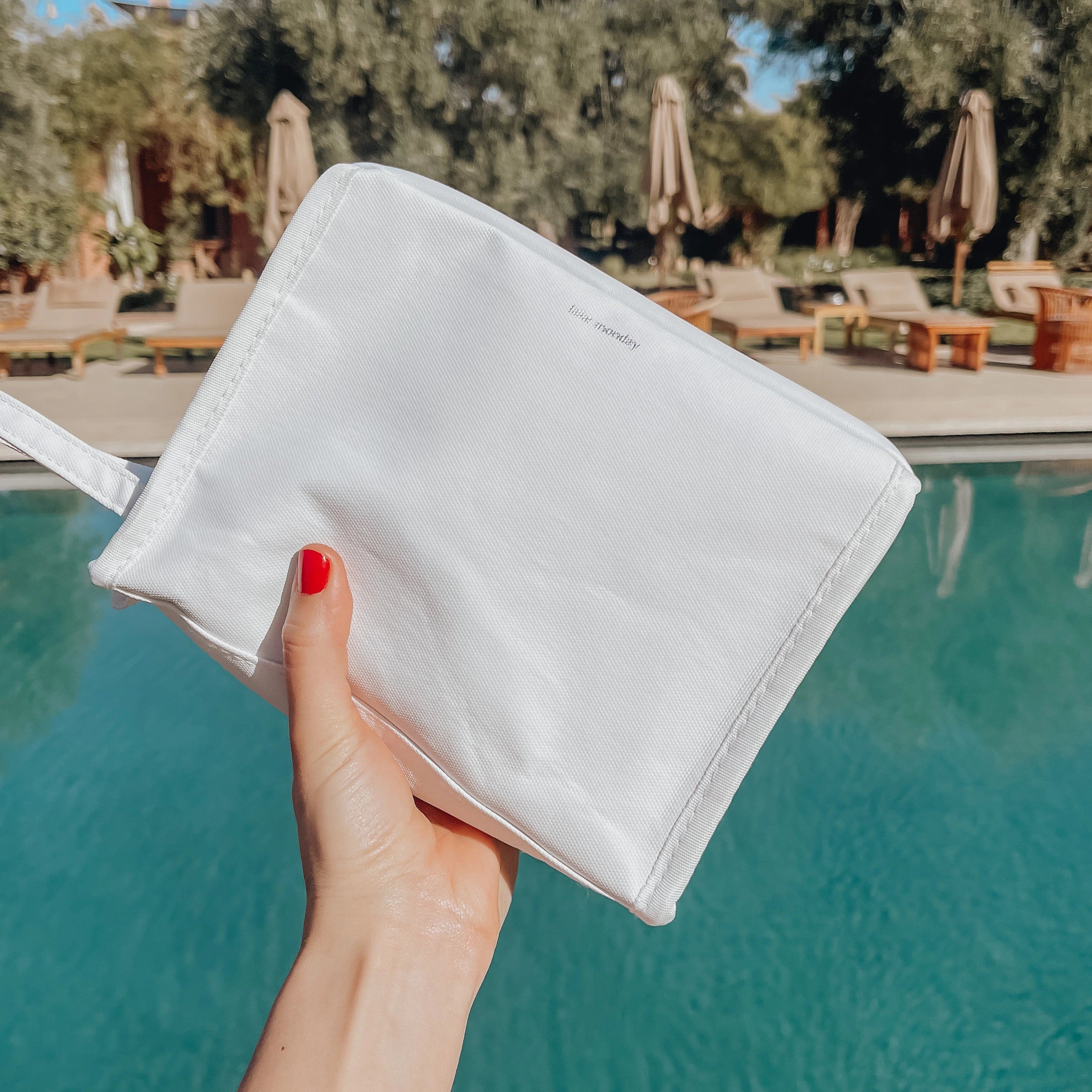 A hand with red nail polish holds a white 'talia moodday' pouch over a vibrant turquoise swimming pool with a sunny resort background featuring lounge chairs and lush greenery