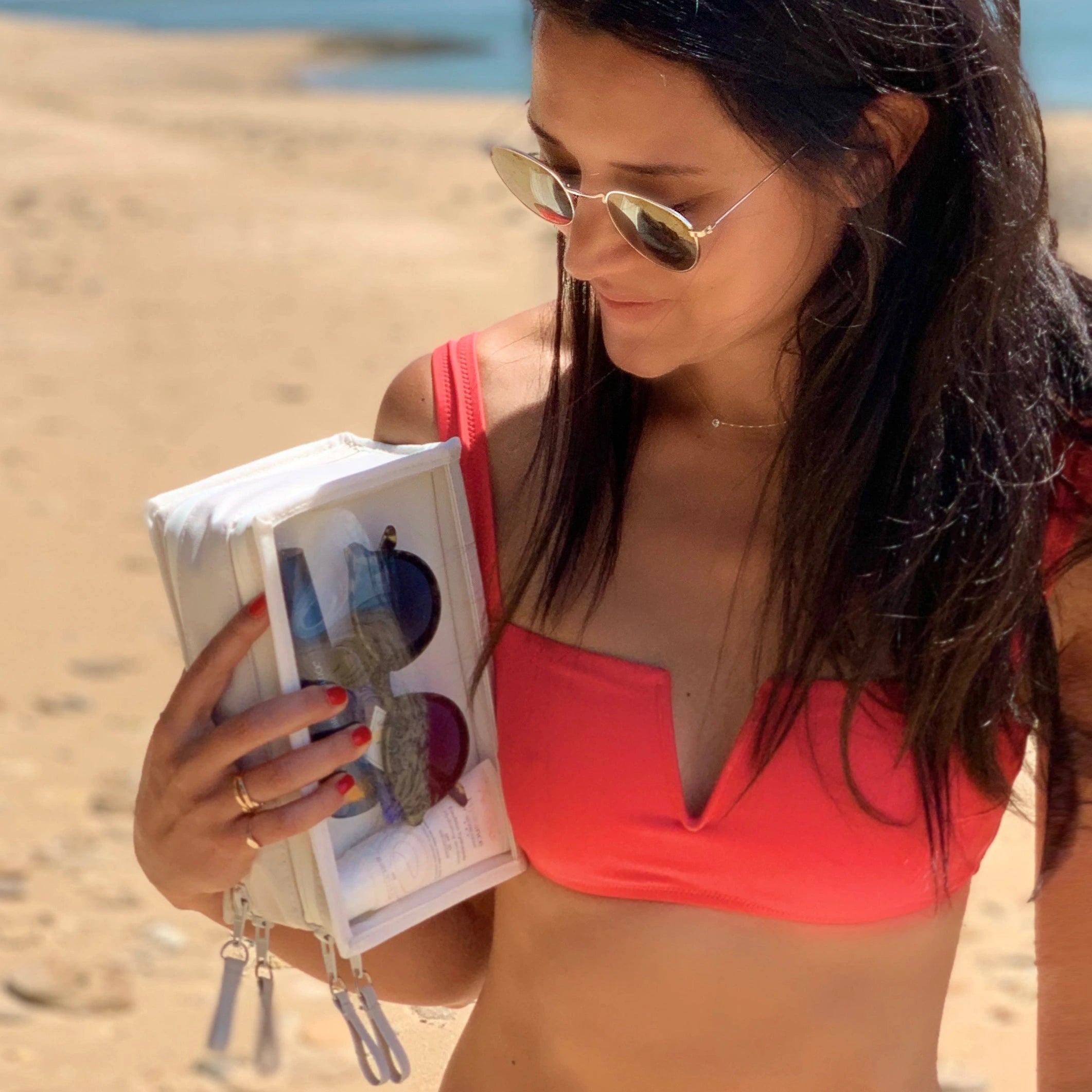 Photograph of a woman wearing a coral bikini top and gold sunglasses holding a clear travel case with sunscreen on a sandy beach