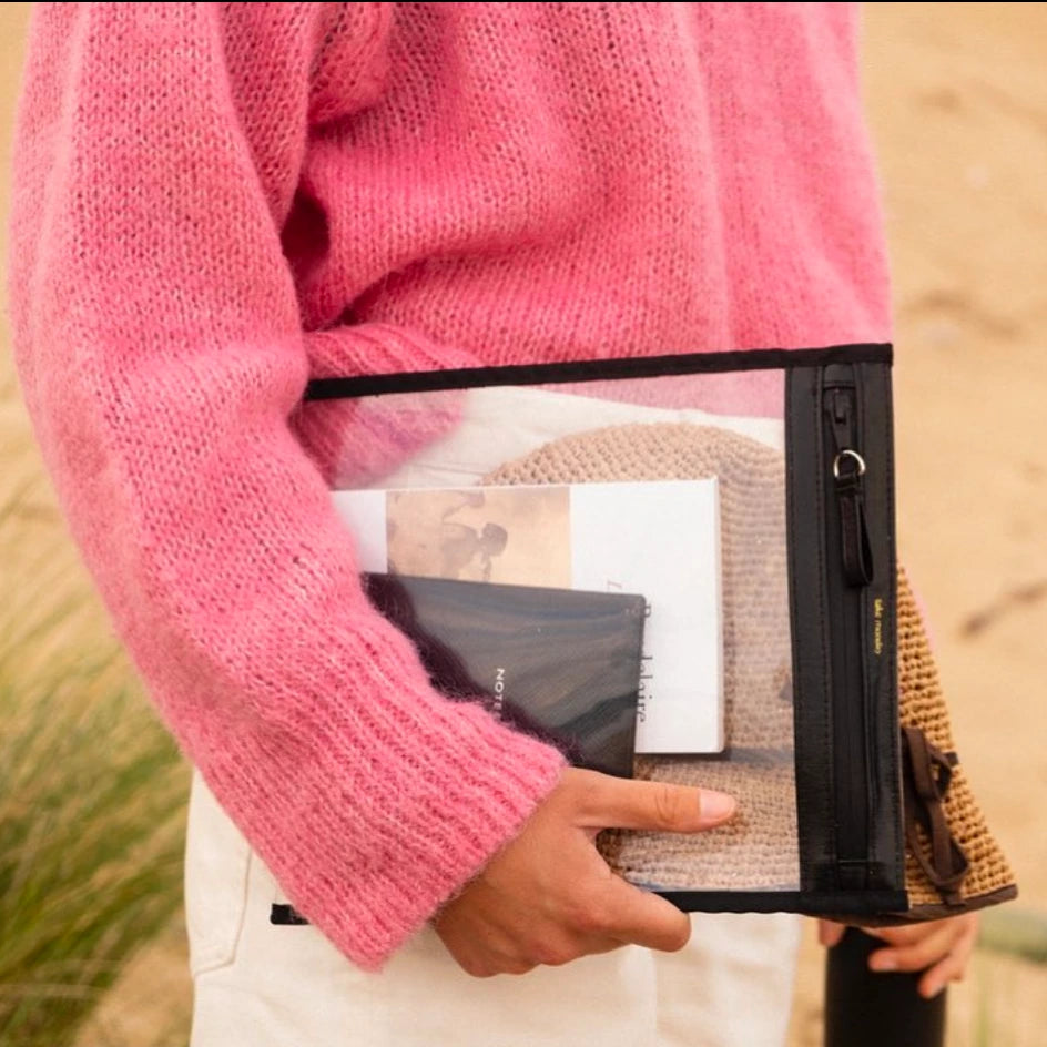 A person in a bright pink fluffy sweater holds a clear PVC pouch containing a beige crocheted hat, a book with a vintage photo, and a dark notebook, against a sandy outdoor background