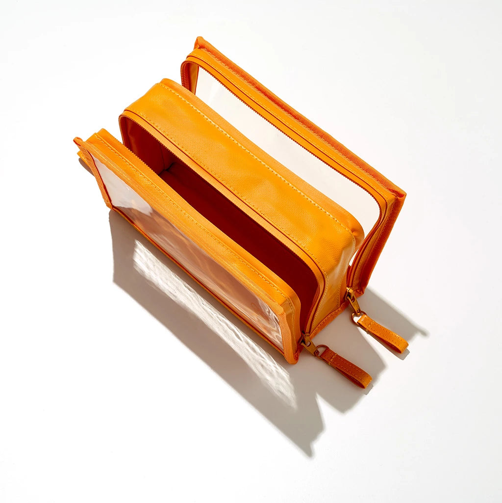 A vibrant orange organizer bag featuring two clear zippered compartments and gold zippers with orange pulls, partially open on a clean white studio background