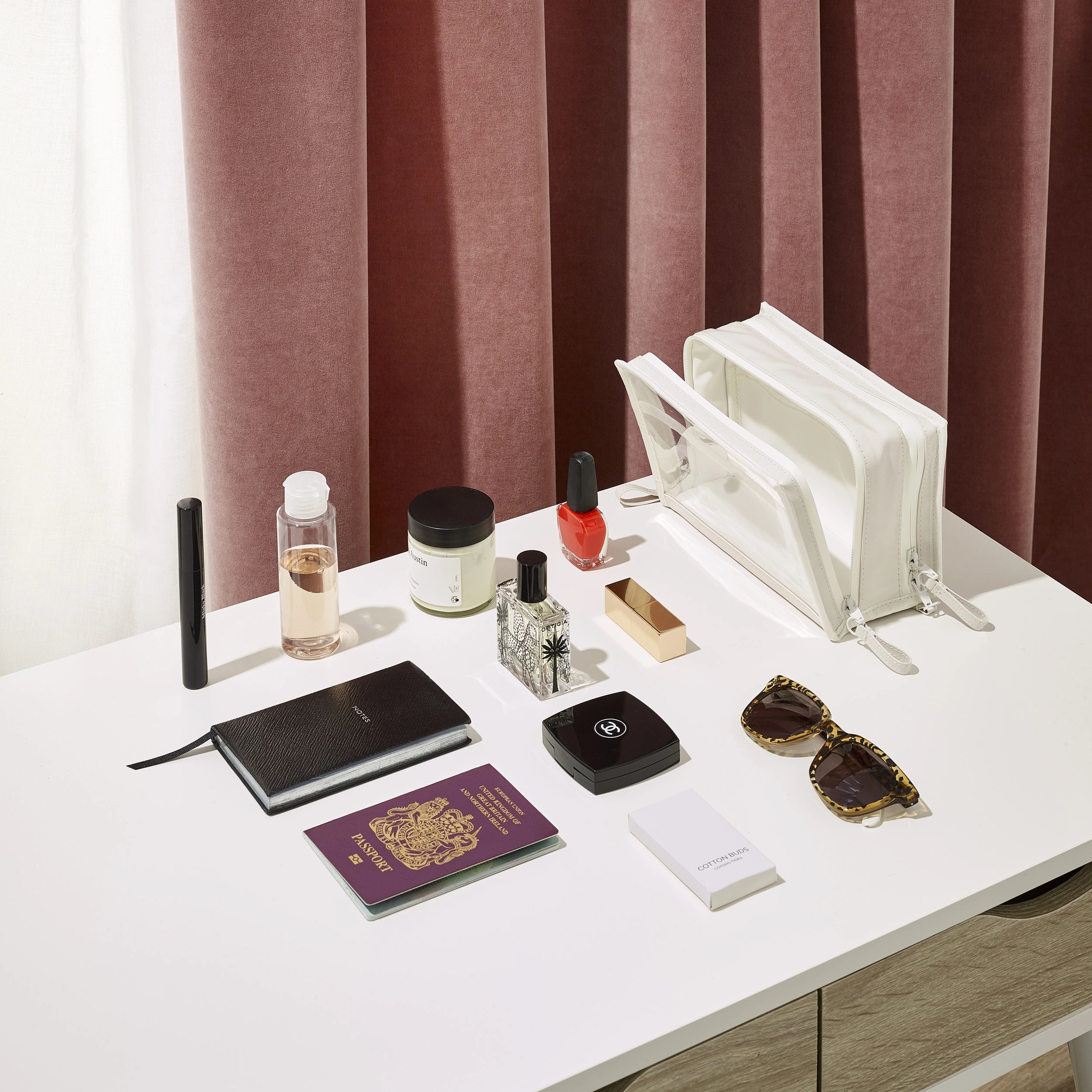 Travel essentials displayed on a white desk in front of plush pink curtains featuring a United Kingdom passport red nail polish and leopard print sunglasses