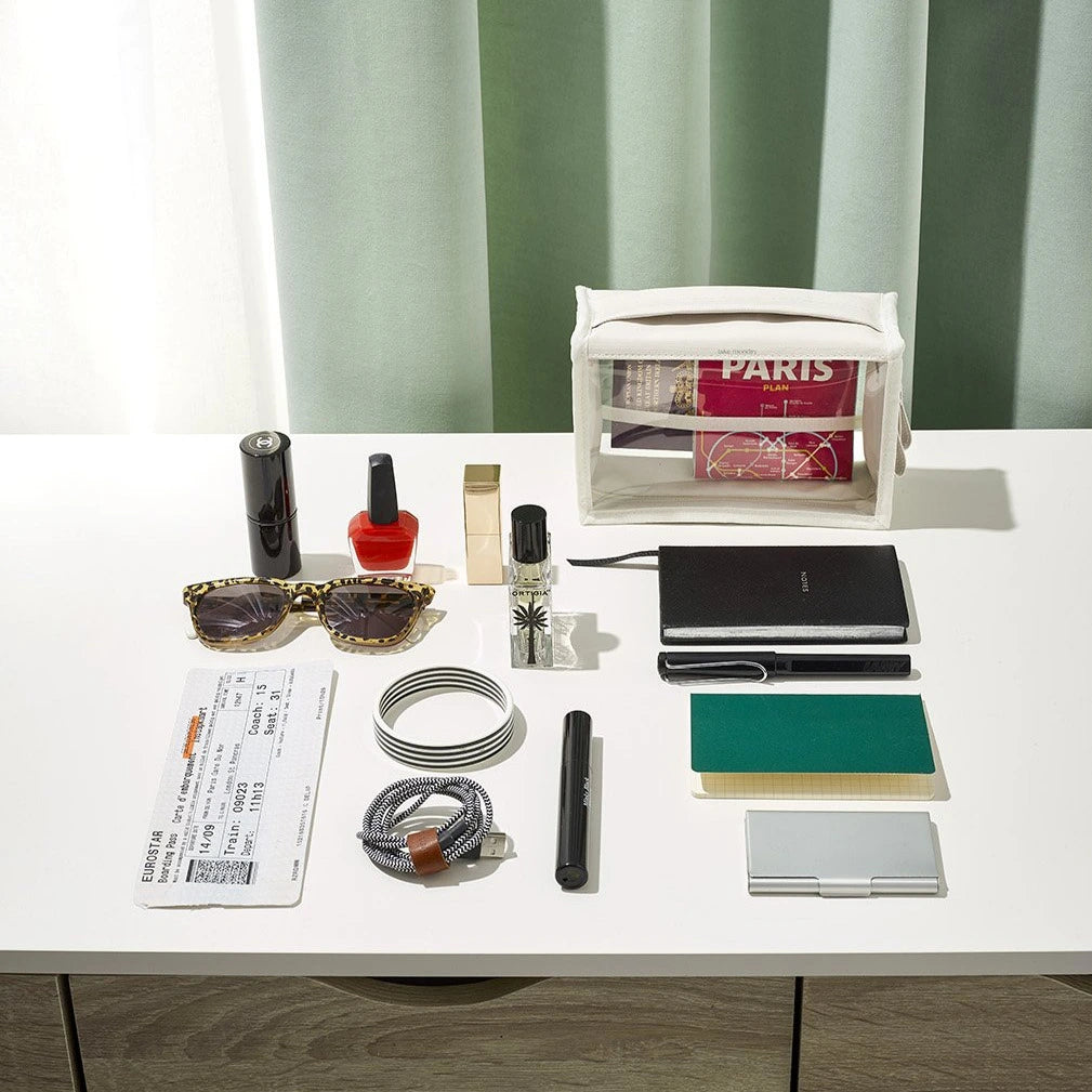 A flat lay of travel essentials for a trip to Paris including a clear pouch with a Paris Plan book, a Eurostar boarding pass, leopard print sunglasses, and red nail polish and lipstick