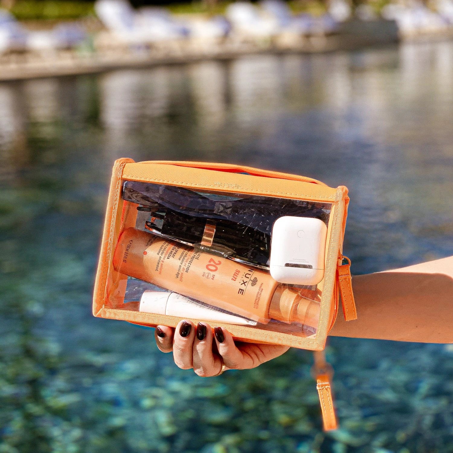 A hand with dark red nail polish holds a transparent orange cosmetic pouch containing Nuxe Sun SPF 20 sunscreen and white AirPods case beside a clear blue resort pool with blurred lounge chairs